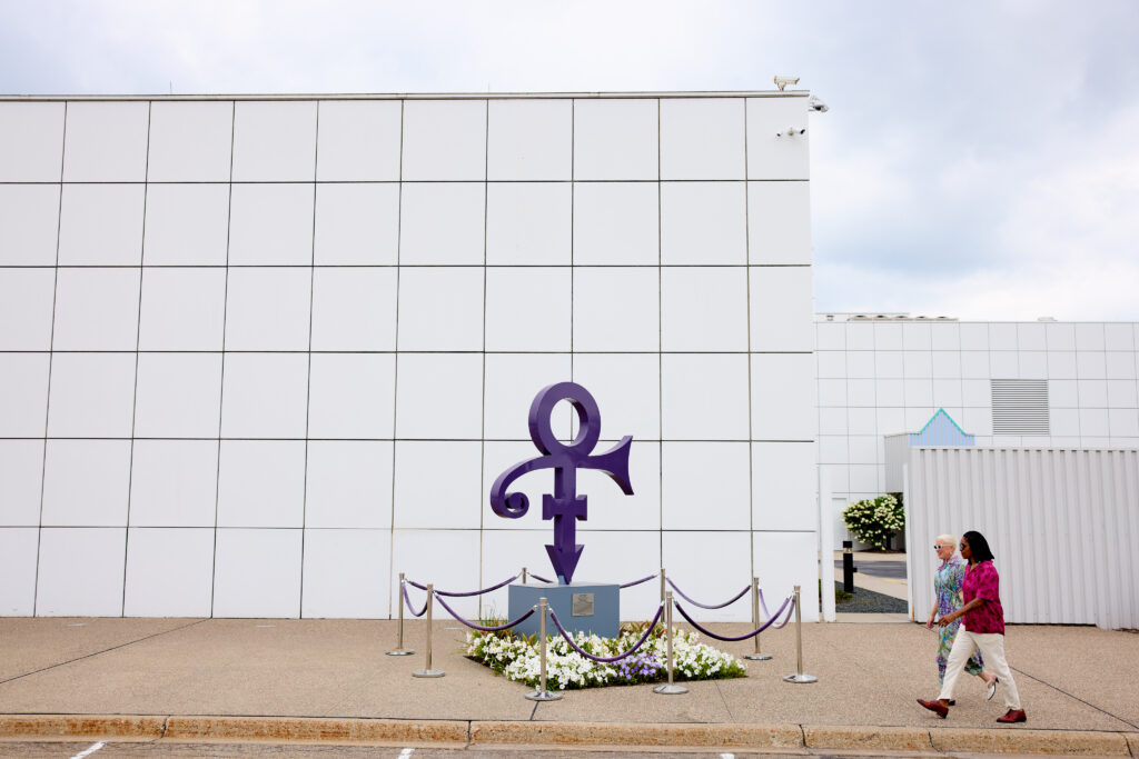 Le symbole violet de l'artiste Prince est présent au centre de l'image, devant le musée Paisley Park. Deux femmes s'avancent vers l'entrée du musée.