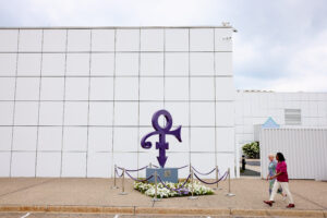 Le symbole violet de l'artiste Prince est présent au centre de l'image, devant le musée Paisley Park. Deux femmes s'avancent vers l'entrée du musée.